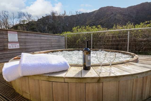 Outdoor wooden hot tub bubbling under a bright sky, with folded white towels, a bottle of wine, and two empty wine glasses placed on the edge, set against a backdrop of rocky hills and spring greenery