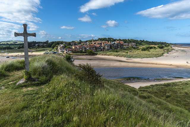 A stone cross standing on a grassy dune overlooking a sandy estuary, with a small coastal village in the background under a bright blue sky with scattered clouds.