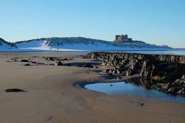 Images shows a lovely winter scene with snow on the dunes with Bamburgh Castle in the distance and an expanse of beach in front of the camera.