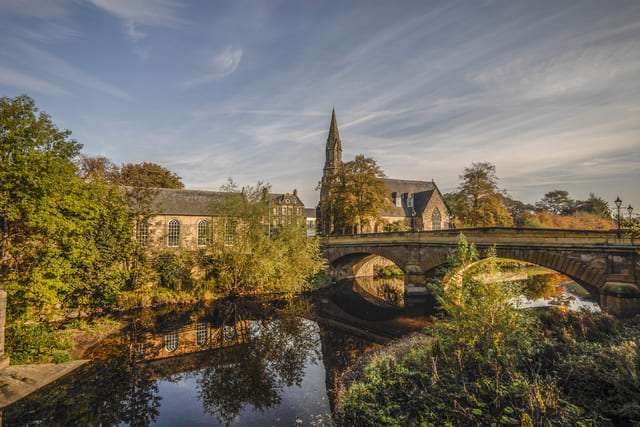 A historic stone church with a tall spire and surrounding autumn trees reflected in a calm river, with an arched stone bridge crossing the water under a soft, golden sky.